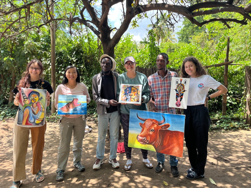 Six people pose, holding colorful artwork in front of bright green shrubbery.