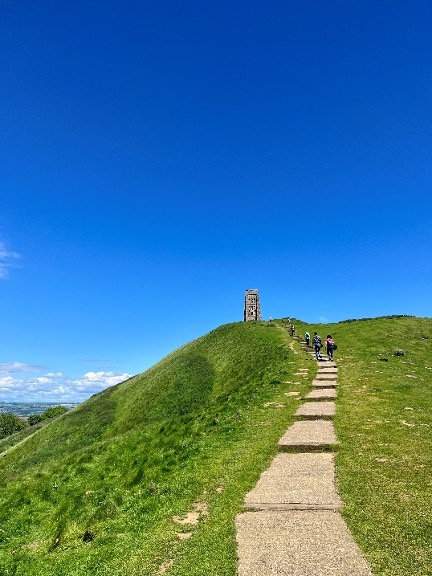Glastonbury Tor sits at the top of a bright green hill. A path leads up to it.