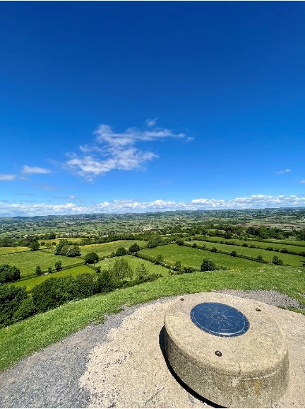The top of Glastonbury Tor overlooks bright green land and a bright blue sky.