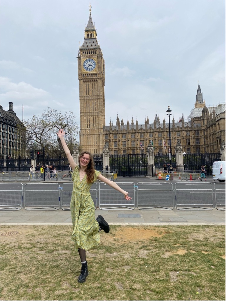 A girl in a long green dress stands in front of Big Ben in London with her arms outstretched.